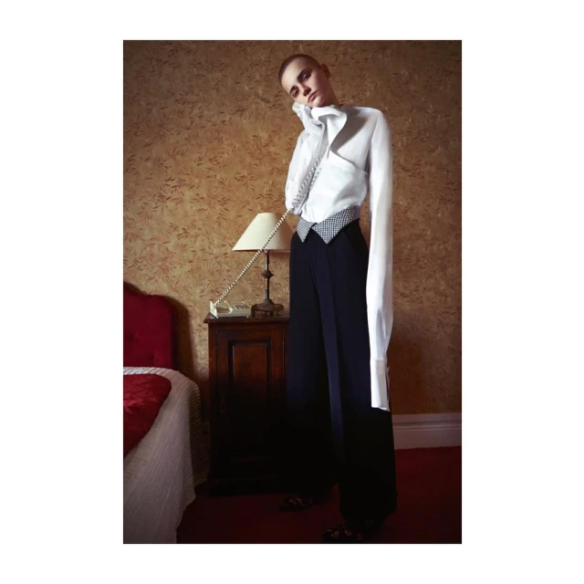 A woman in a white shirt and dark pants poses with a vintage telephone in a room with a bed, creating a fashion editorial image.