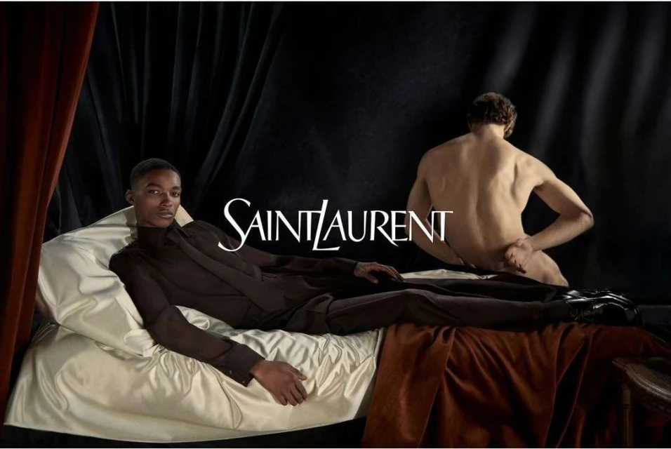 Fashion editorial shot featuring an African American man in a dark suit reclining on a bed, with Saint Laurent logo text overlay.