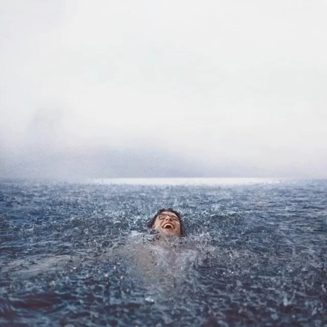 Adult male swims and laughs in a body of water in the daytime under an overcast sky, looking up. Waves and rain surround him.