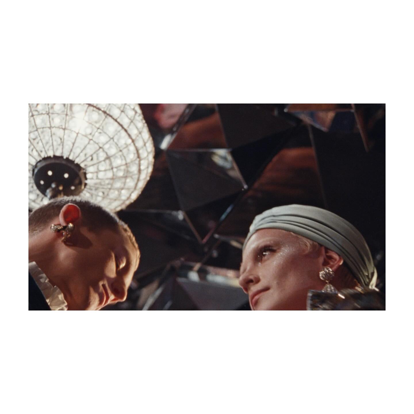 Close-up of two adults in a mirrored room with a chandelier. One person wears a head covering, the other has a short haircut.