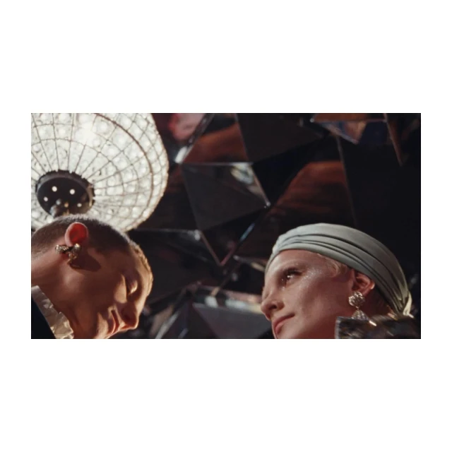 Close-up of two adults in a mirrored room with a chandelier. One person wears a head covering, the other has a short haircut.