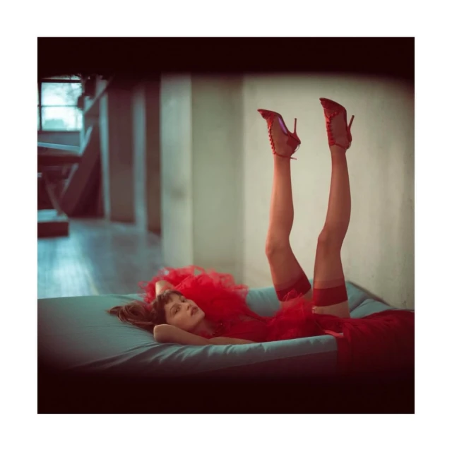 Brunette woman in a red dress lies on a blue surface with her legs raised, wearing red heels and red stockings against a neutral background.