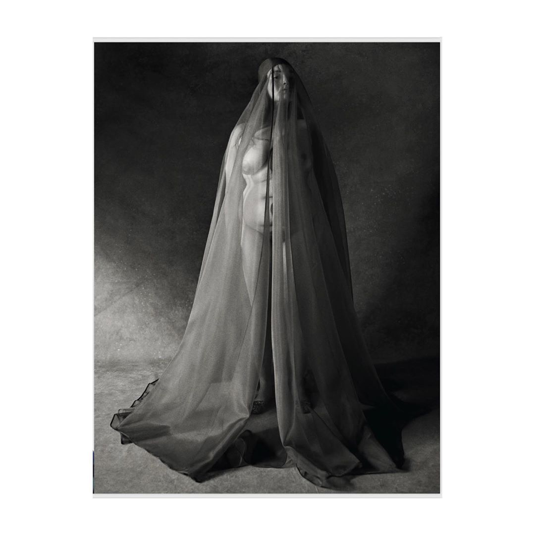 Monochrome portrait of a nude woman standing indoors, draped in a sheer veil from head to toe against a dark grey backdrop.