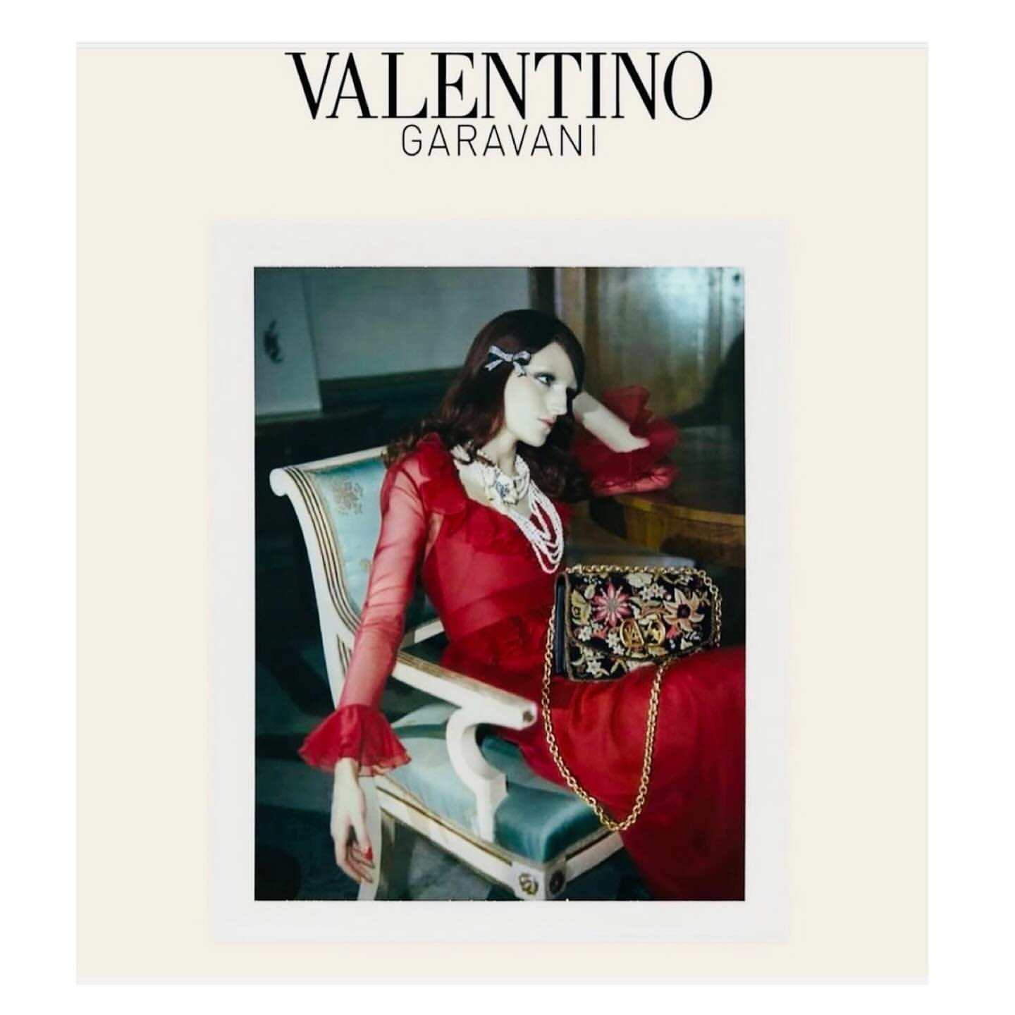 Elegant mannequin seated in chair, adorned in red gown and pearl jewelry, with a floral purse in a vintage-inspired fashion photo.