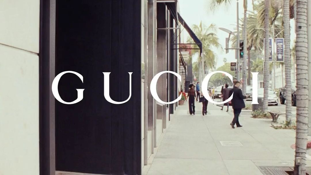 Street scene with Gucci store signage, pedestrians walking on the sidewalk, and palm trees lining the road on a sunny day.