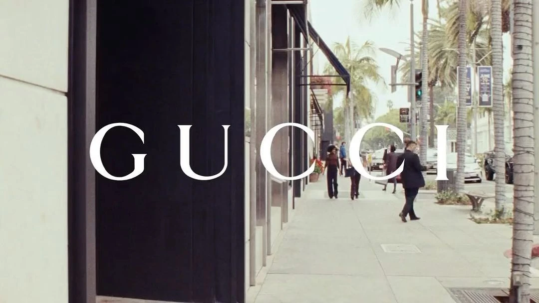 Street scene with Gucci store signage, pedestrians walking on the sidewalk, and palm trees lining the road on a sunny day.