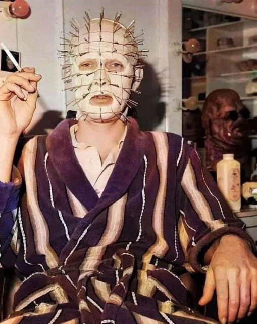Man in striped robe with nails in head smokes cigarette indoors, grotesque mask visible in the background. Horror and celebrity. 