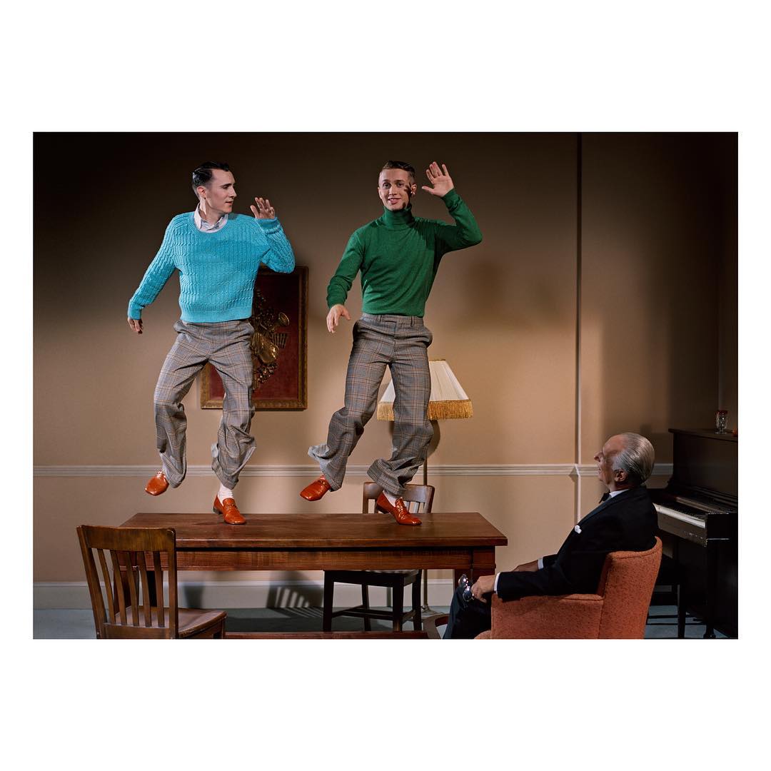 Two men in sweaters and trousers jump on a desk, while an older man in a suit sits in a chair and watches in an indoor setting.