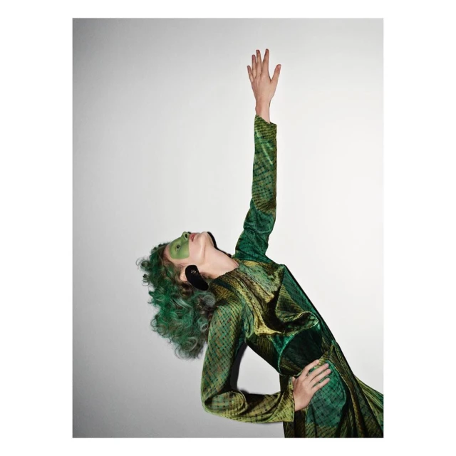 A woman in a patterned green dress reaches upward with green makeup. The portrait is shot in a studio against a gradient background.
