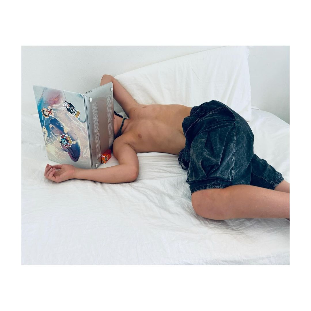 Teenage male lying on a white bed, head resting on a closed laptop computer with cartoon graphics, studio lighting, casual posture.