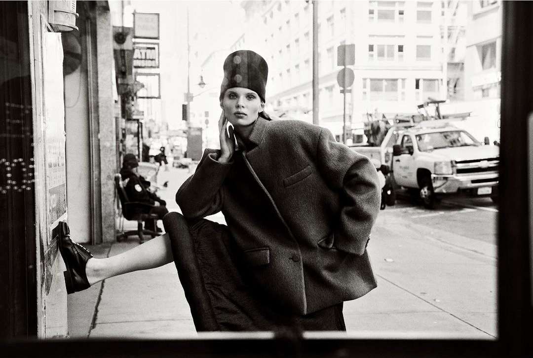 Monochrome portrait of a woman in a coat and hat posing on a city sidewalk with buildings and a truck in the background.