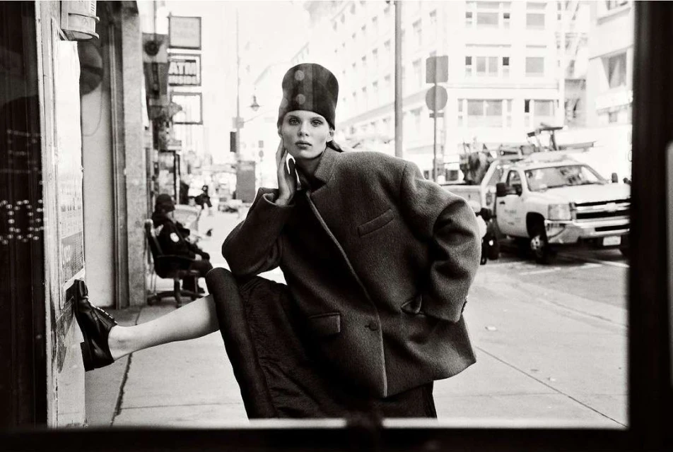Monochrome portrait of a woman in a coat and hat posing on a city sidewalk with buildings and a truck in the background.