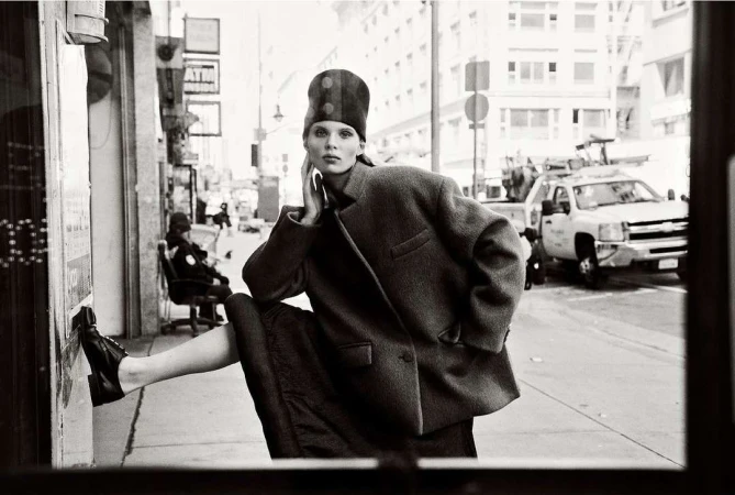 Monochrome portrait of a woman in a coat and hat posing on a city sidewalk with buildings and a truck in the background.