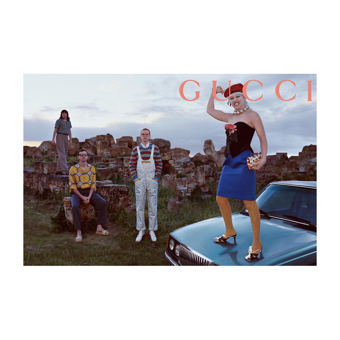 Fashion editorial featuring a group of five adults in an outdoor setting near a car and stone ruins under a cloudy sky at dusk.