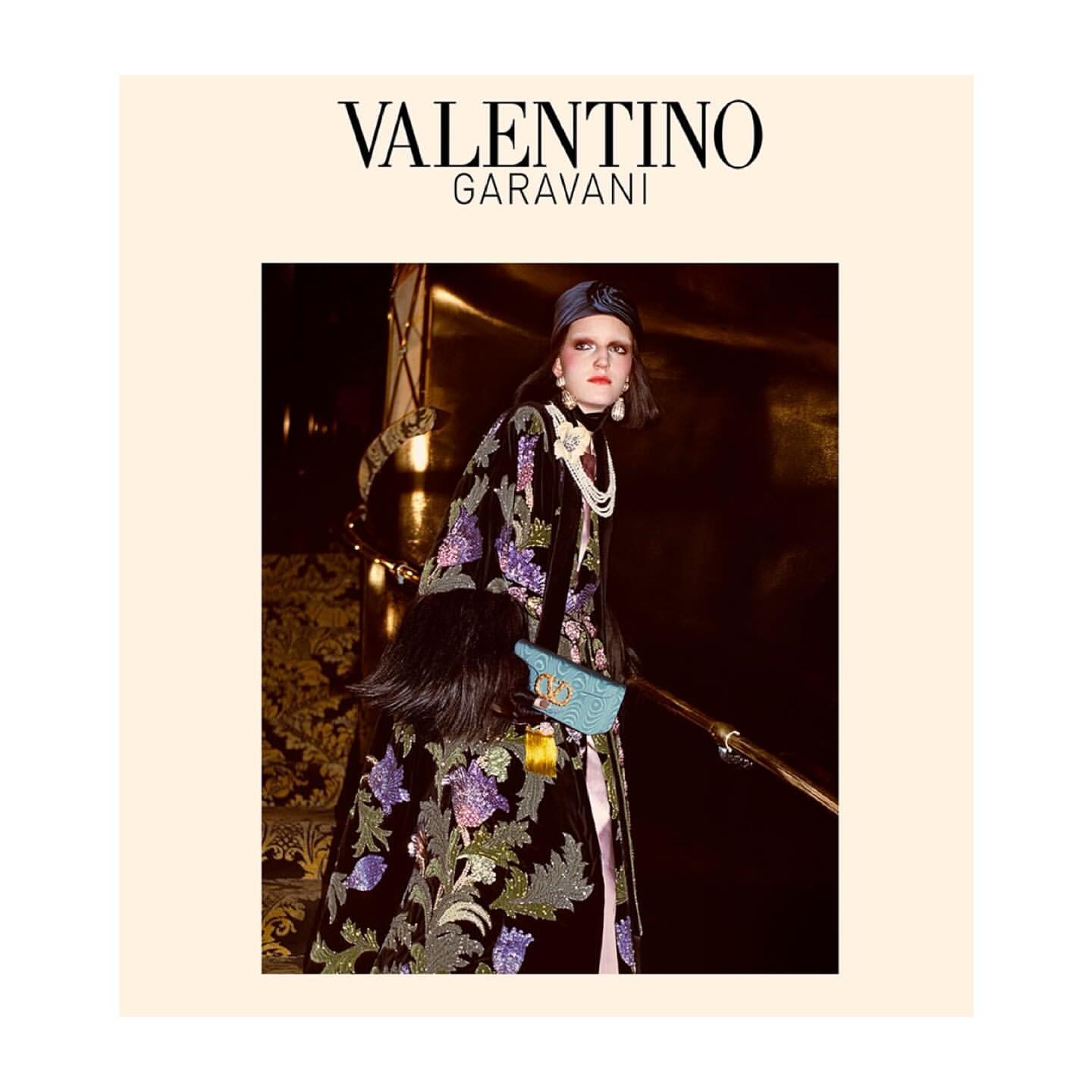Fashion photography featuring a stylish adult female model in a floral coat and clutch, posing against a golden interior backdrop.