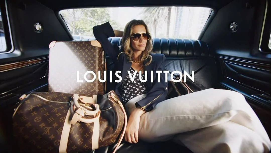 A woman in sunglasses lounges in a luxury car surrounded by Louis Vuitton luggage. The interior is sleek and the scene is well-lit.