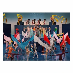Wide ensemble shot of performers in colourful theatrical costumes holding large flags on a tiered blue stage set by Glen Luchford
