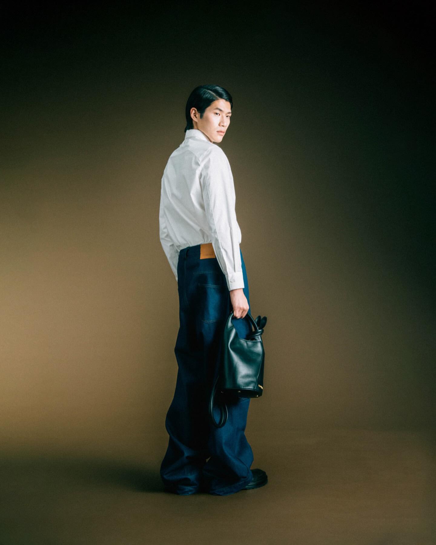 Full shot of a young adult male in a white button-down and baggy jeans holding a small black bag against a neutral gradient backdrop.