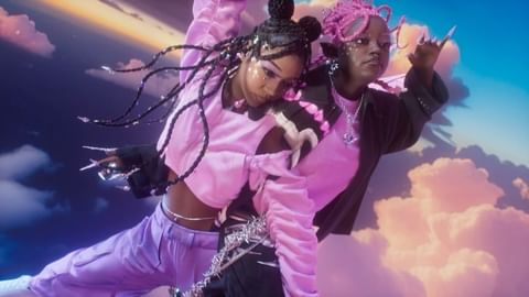 Two stylish young women pose in front of a dreamy cloudy sky. One wears purple and the other wears black and pink. They have long, decorated fingernails.