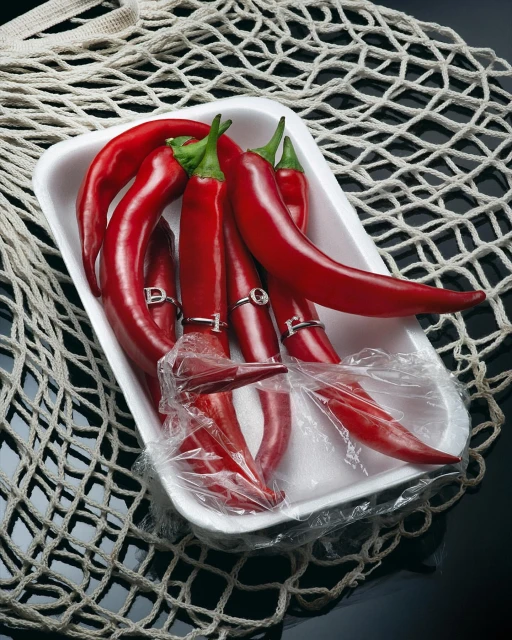 Artistic food still life: Fresh red chili peppers are displayed in a white tray with decorative rings, against a net and dark backdrop.