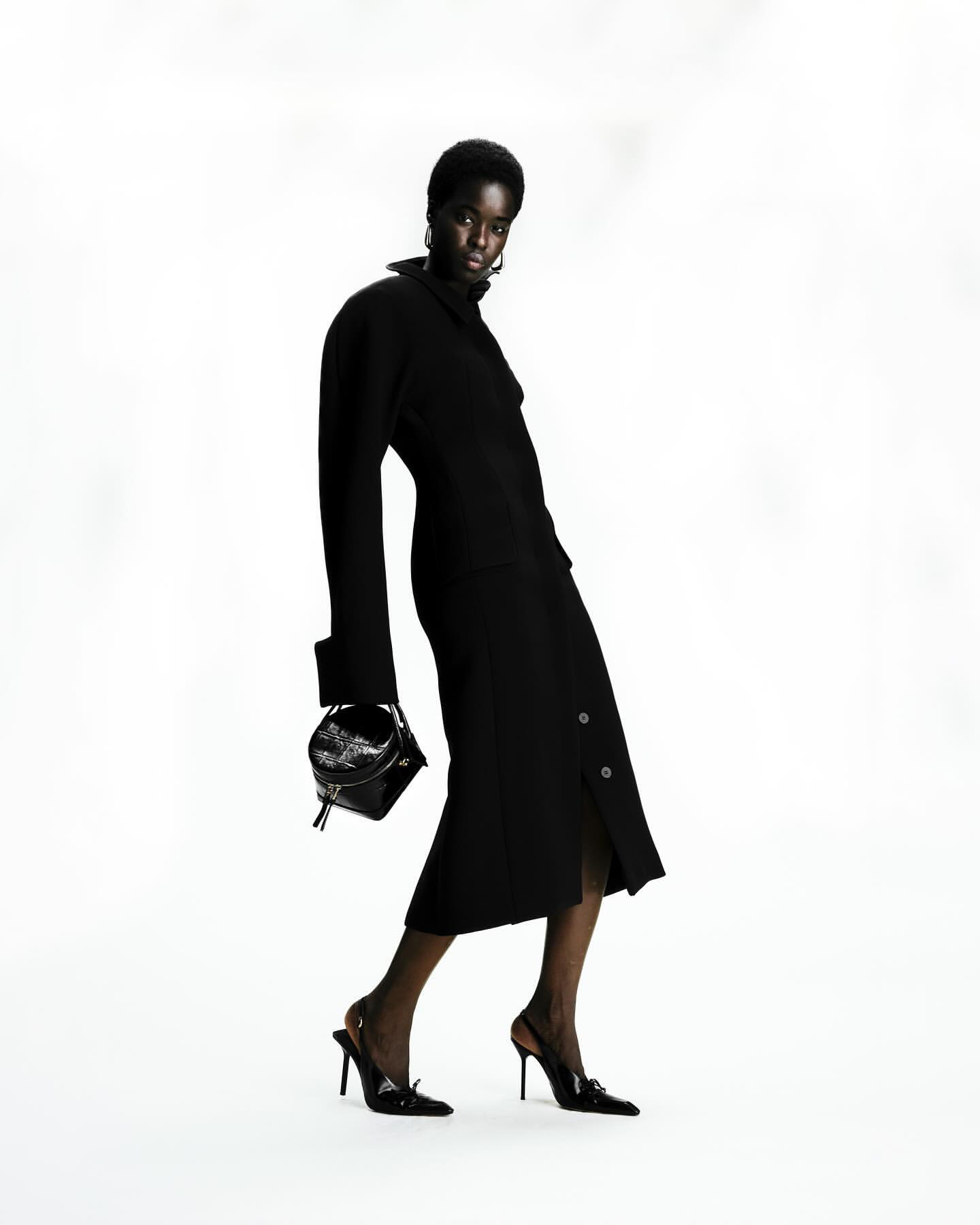 Dark-skinned female fashion model in a black dress and black heels, carrying a structured black purse against a white backdrop.