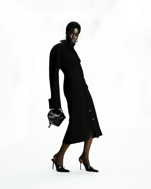Dark-skinned female fashion model in a black dress and black heels, carrying a structured black purse against a white backdrop.