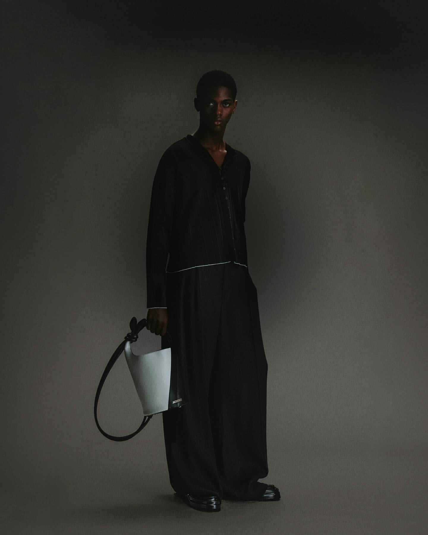 Portrait of a Black male model in a black outfit holding a light grey handbag in a studio with a dark grey background.
