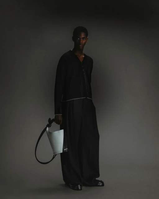 Portrait of a Black male model in a black outfit holding a light grey handbag in a studio with a dark grey background.
