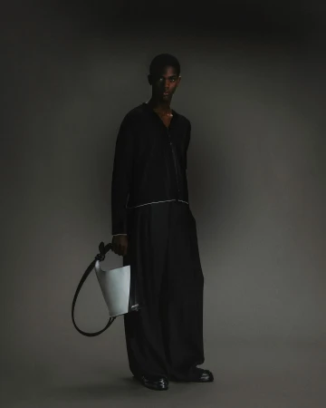 Portrait of a Black male model in a black outfit holding a light grey handbag in a studio with a dark grey background.