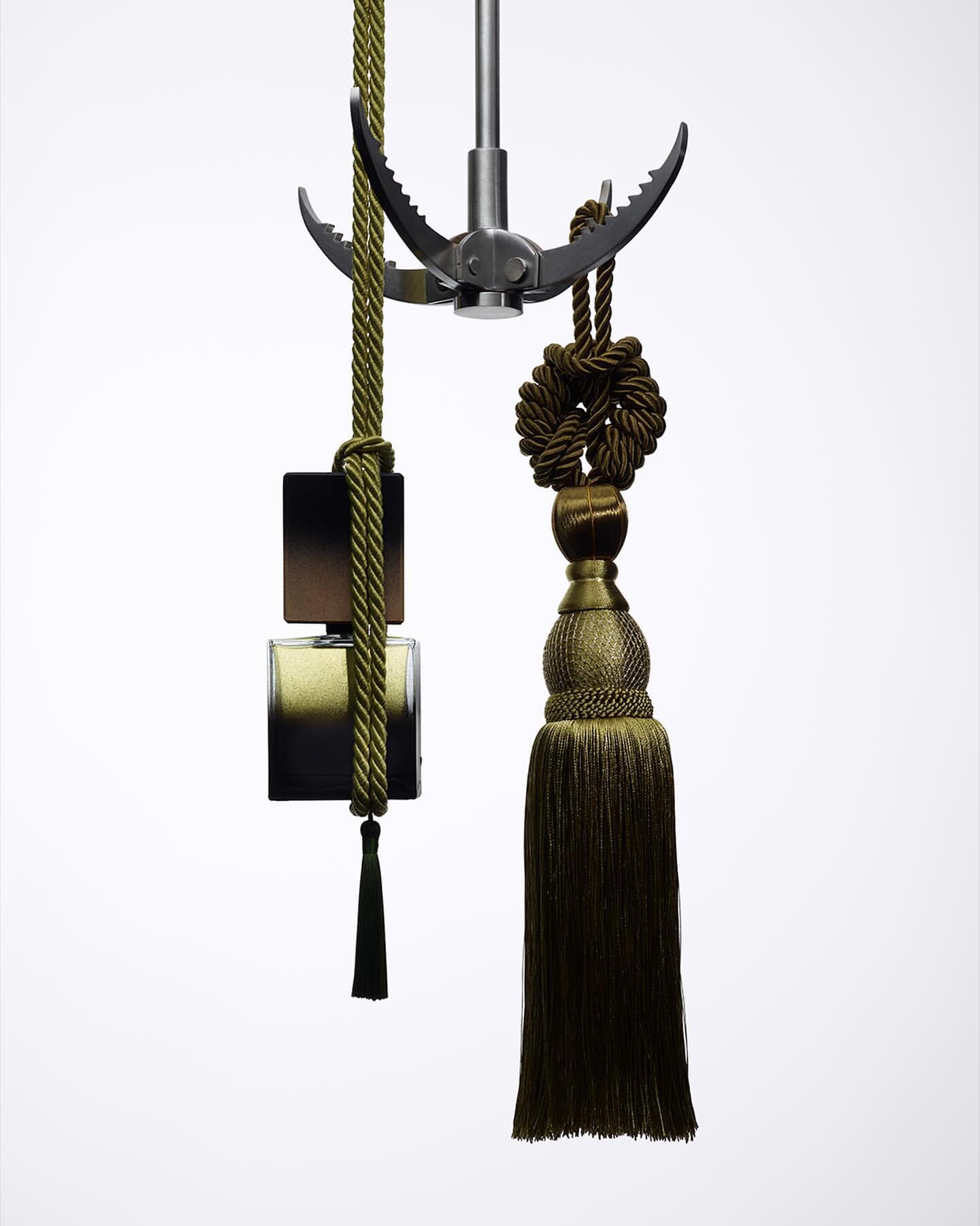 Conceptual display of a dark-toned perfume bottle suspended by decorative ropes and a metallic grappling hook against a bright white background.
