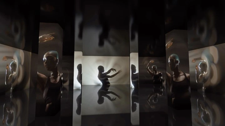 Person in a dark tank top stands against a bright white wall, reflected in dark mirror panels, creating an illusion of multiple figures.