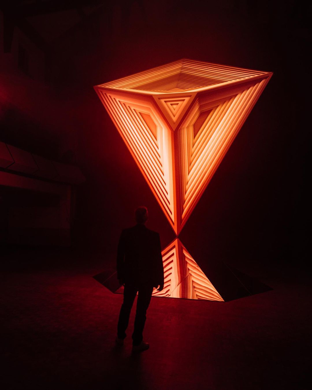 Person in dark space looks up at layered geometric structure illuminated in orange light with reflection on floor, creating dramatic effect.