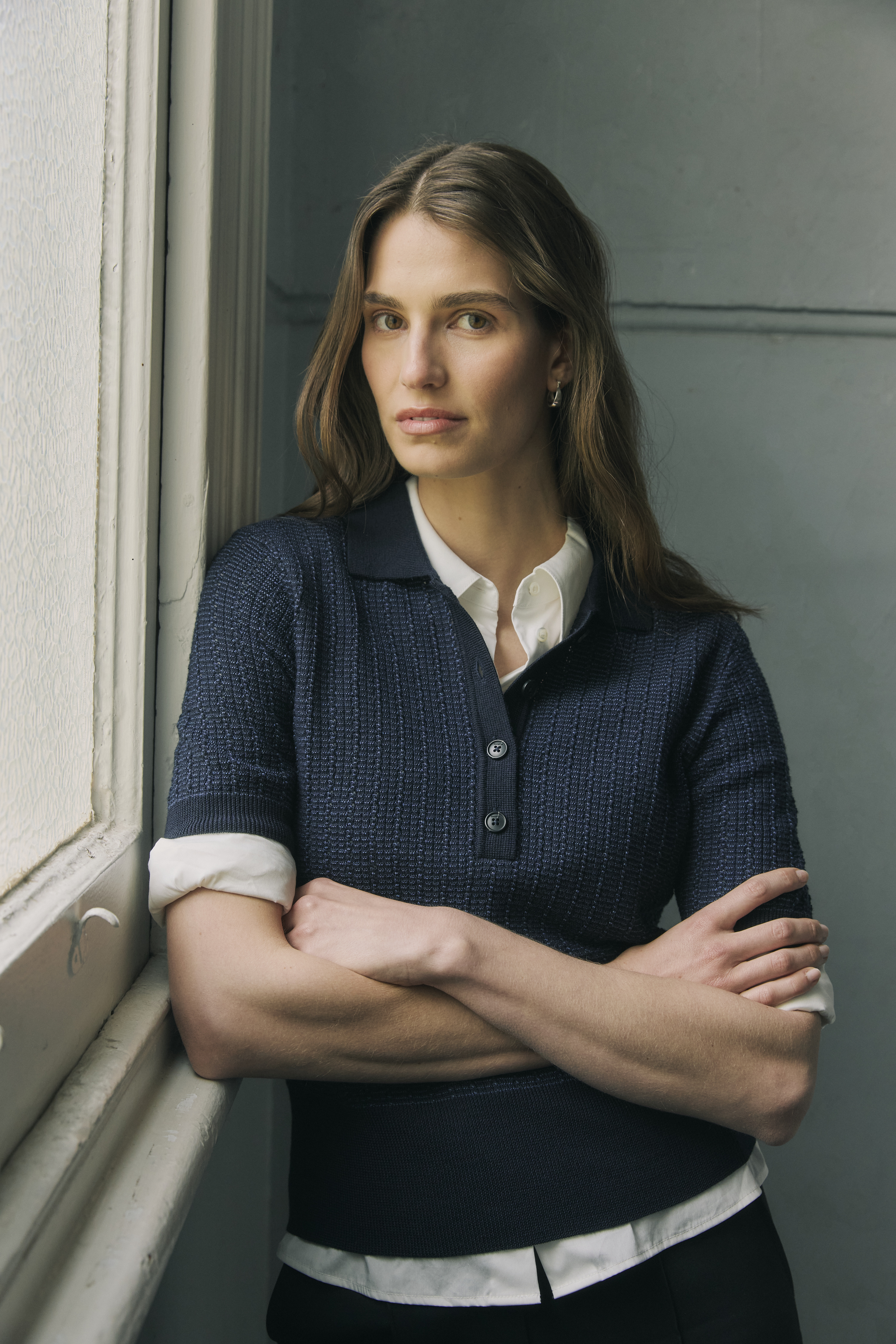 Portrait of a woman with arms crossed, wearing a navy collared knit shirt over a white collared shirt, standing next to a window.
