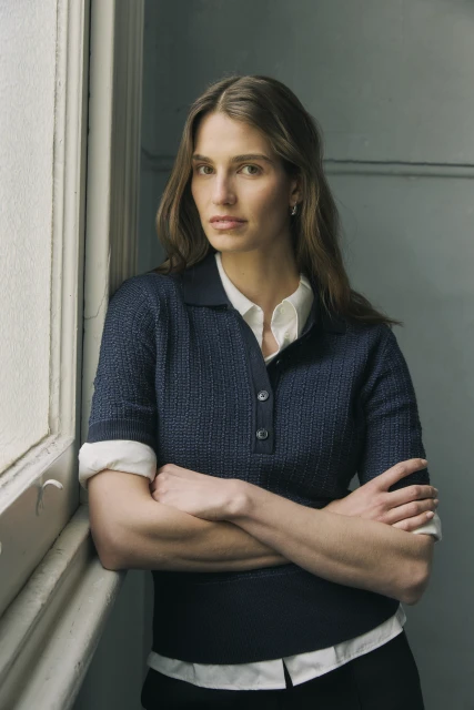 Portrait of a woman with arms crossed, wearing a navy collared knit shirt over a white collared shirt, standing next to a window.