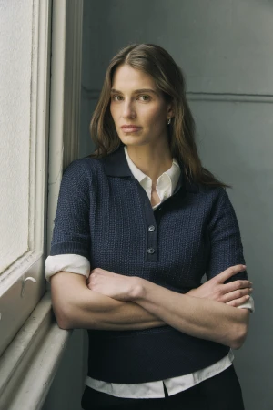 Portrait of a woman with arms crossed, wearing a navy collared knit shirt over a white collared shirt, standing next to a window.