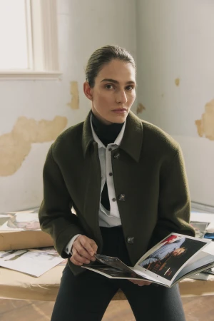 Woman in an olive green wool jacket holding a magazine seated indoors by a window in a portrait by photographer Bella Dower.