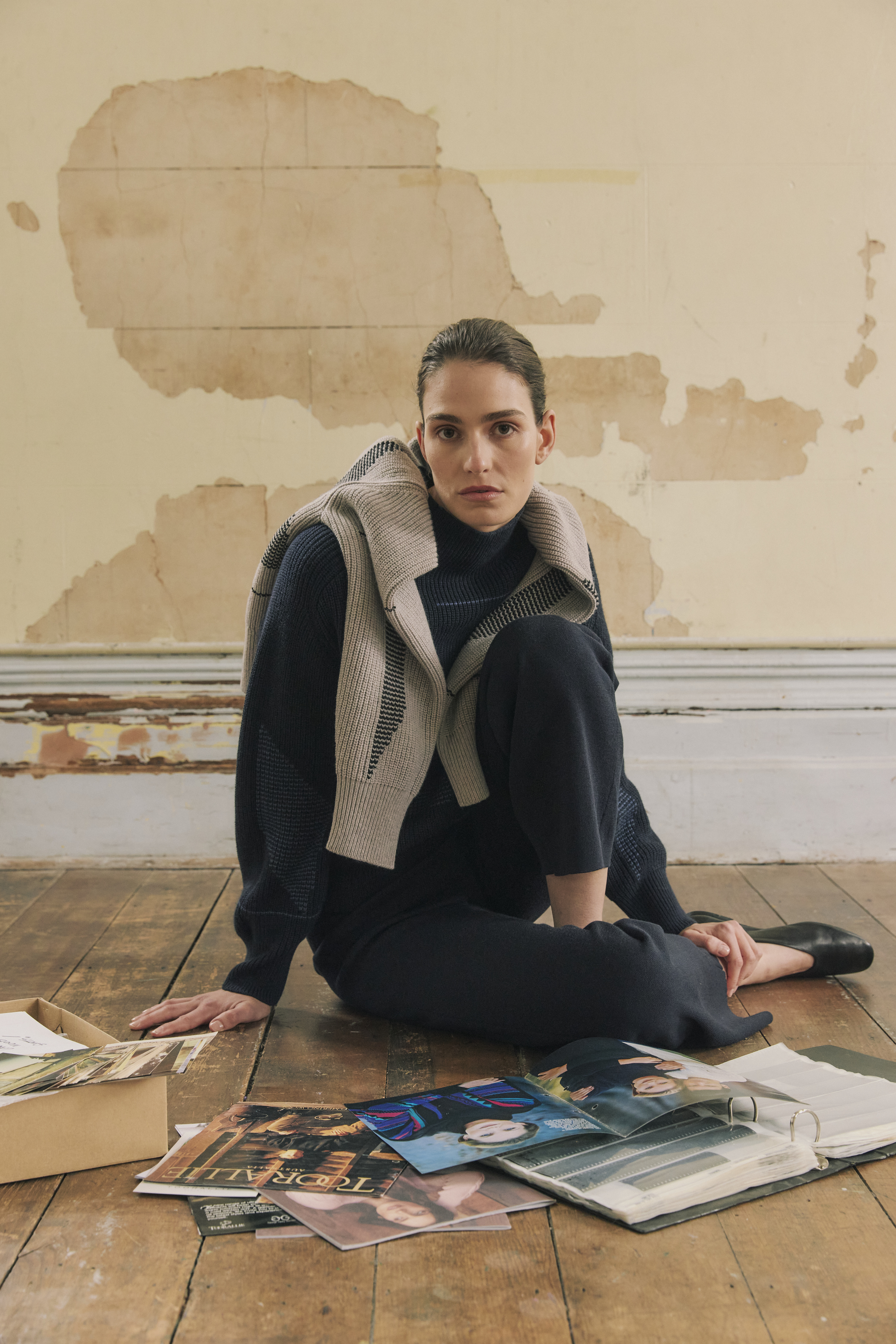 A woman in a navy outfit poses in a worn indoor studio space with aged wall paint, seated on a wooden floor with old photos spread around.