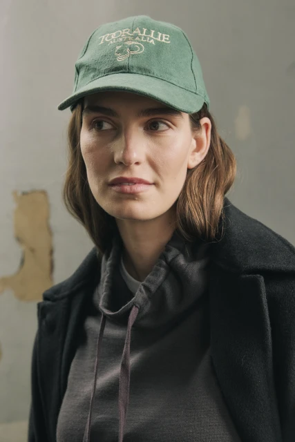 Woman wears a green baseball cap indoors, paired with a gray hoodie and a dark coat, posing against a textured gray wall backdrop.