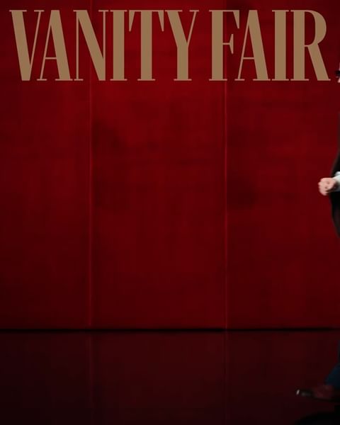 Person in dark suit stands in front of a red wall with VANITY FAIR text above.
