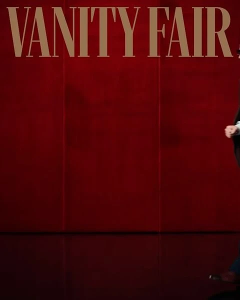 Person in dark suit stands in front of a red wall with VANITY FAIR text above. 