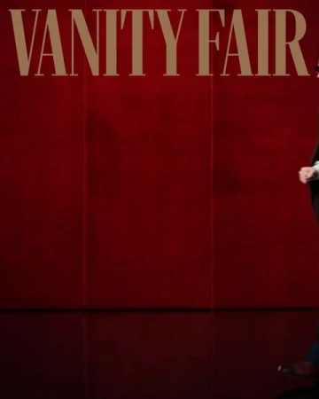 Person in dark suit stands in front of a red wall with VANITY FAIR text above. 