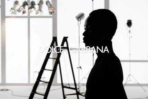 Black and white studio shot of a silhouetted figure and ladders, with lighting equipment and a Dolce & Gabbana logo.
