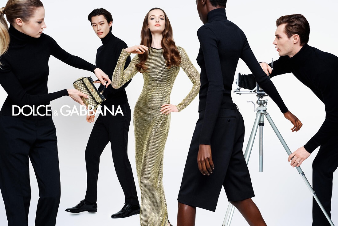 Fashion editorial featuring five diverse models wearing black turtlenecks and a gold dress, holding a gold handbag and old-fashioned camera.