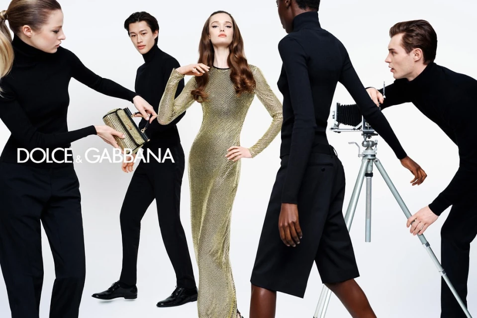 Fashion editorial featuring five diverse models wearing black turtlenecks and a gold dress, holding a gold handbag and old-fashioned camera.