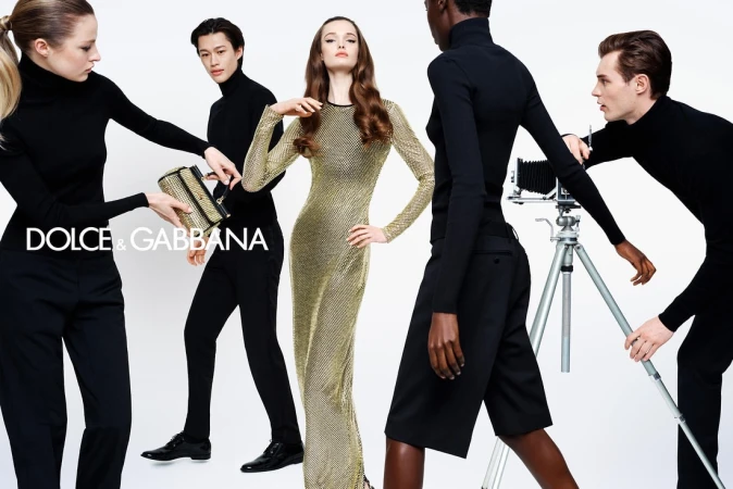 Fashion editorial featuring five diverse models wearing black turtlenecks and a gold dress, holding a gold handbag and old-fashioned camera.