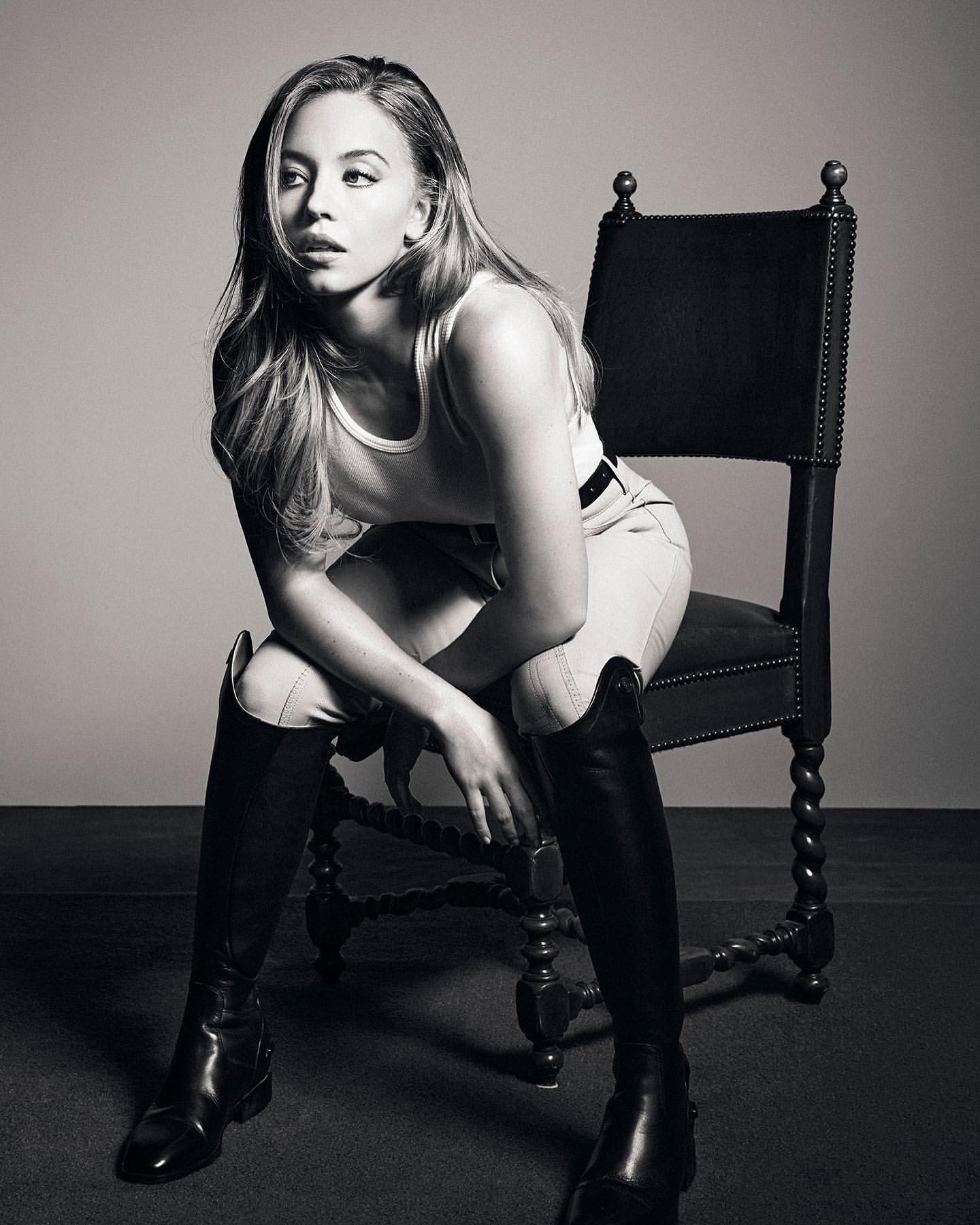 Black and white portrait of a young adult woman with long hair posing while sitting on an antique chair with knee-high boots.