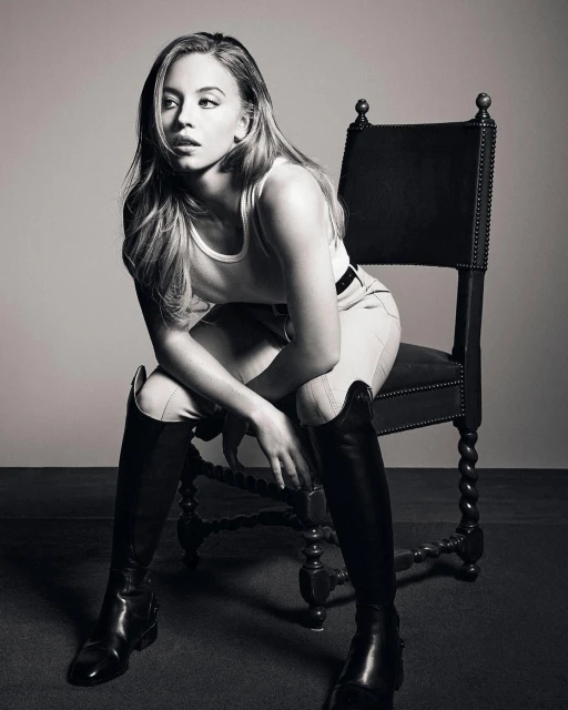 Black and white portrait of a young adult woman with long hair posing while sitting on an antique chair with knee-high boots.