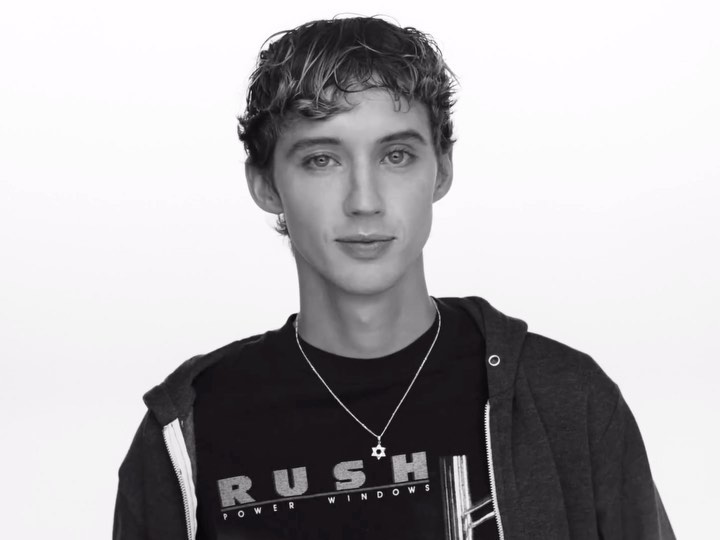 Black and white portrait of a young adult male with curly hair wearing a hooded sweatshirt and t-shirt against a white backdrop.