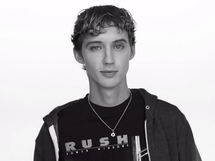 Black and white portrait of a young adult male with curly hair wearing a hooded sweatshirt and t-shirt against a white backdrop.