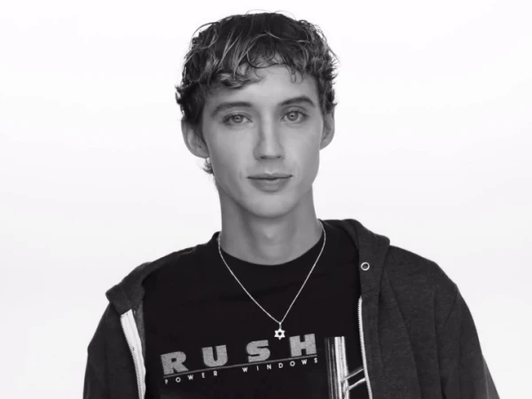 Black and white portrait of a young adult male with curly hair wearing a hooded sweatshirt and t-shirt against a white backdrop.